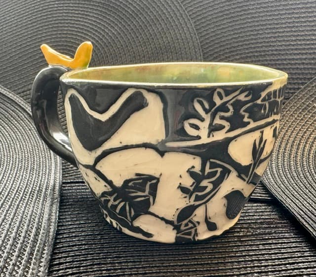 Handmade ceramic mug with black and white nature patterns and a yellow bird handle.