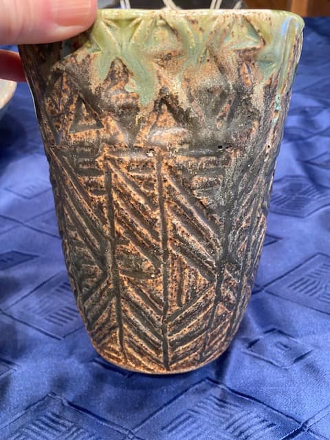 Textured ceramic vessel with carved geometric patterns and green glaze against blue patterned fabric.