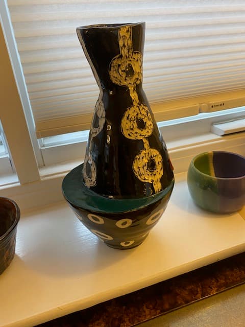 Black ceramic vase with white circular patterns resting on a white windowsill near a window.