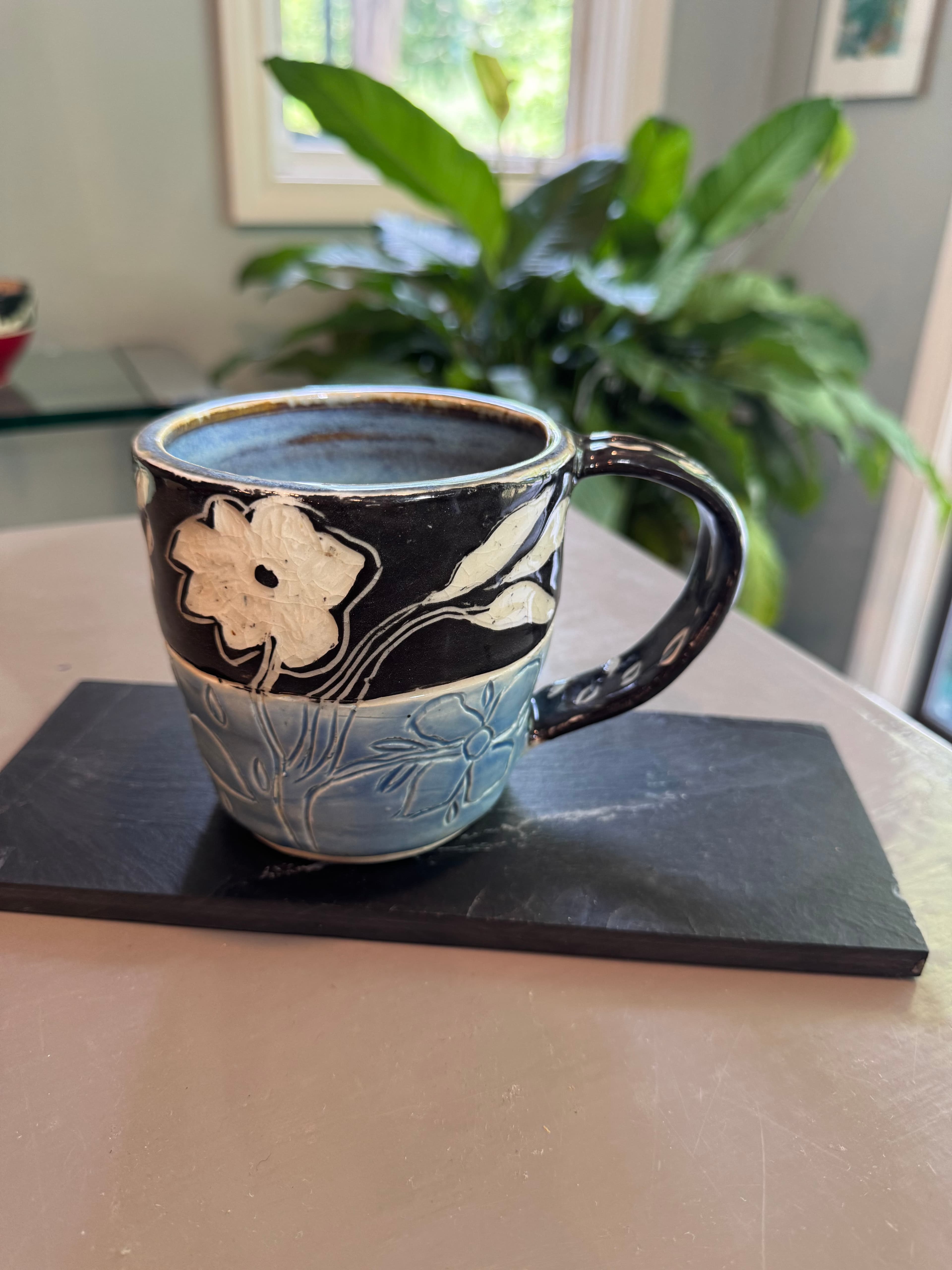 Handcrafted ceramic mug with white floral patterns on black and light blue glazes, on slate.